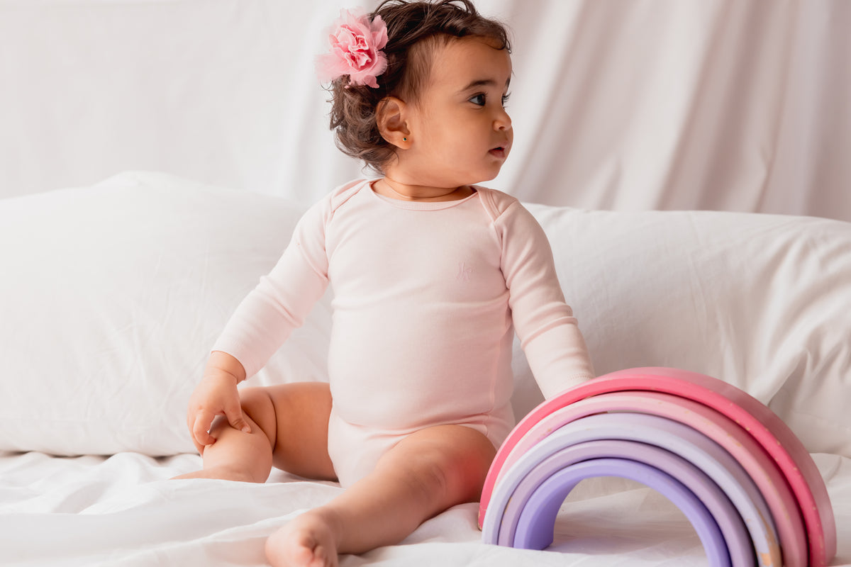 Baby sitting in a cream long sleeve bodysuit beside a rainbow toy
